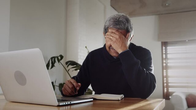 Multi-cultural elderly male upset, recieving bad news while on laptop, sitting at kitchen counter