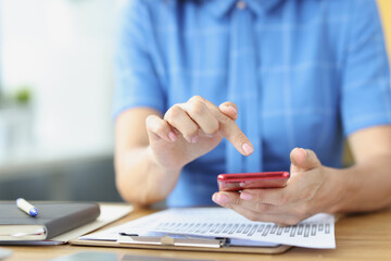 Female finger pressing on smartphone screen over documents with graphs in office closeup