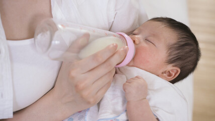 young infant is drinking milk with eyes close while its parent is using fingers to touch the bottle lightly in the bedchamber with cropped shot.