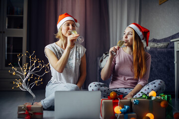 Two happy excited young women in Santa Claus Christmas hats drinking champagne sitting on the floor in a cozy home interior among gifts and burning garlands.