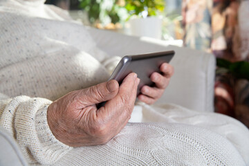 Close-up wrinkled hands of old woman with tablet computer. Senior lady sitting in chair with blanket. Warm and cozy, time to relax, browsing, watching movie. 