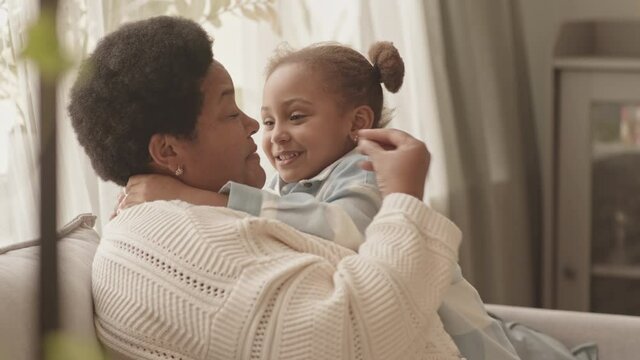 Medium close-up of joyful little girl hugging and kissing her favorite African American granny, sitting on couch in apartment at daytime