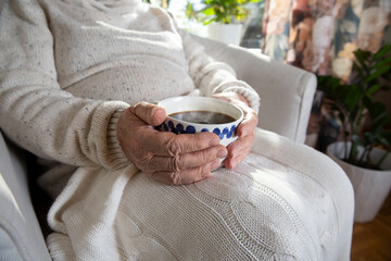 Close-up hands of old woman with mug of hot coffee. Senior lady sitting in chair with blanket. Warm...