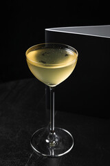 A close-up of an alcoholic cocktail in a nick and nora glass garnished with black sesame seeds and oil drops, a menu book on black wooden table, black background