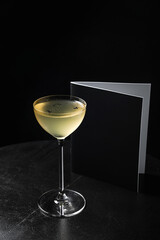 An alcoholic cocktail in a nick and nora glass garnished with black sesame seeds and oil drops, a menu book on black wooden table, black background