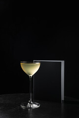 An alcoholic cocktail in a nick and nora glass and a menu book on black wooden table, black background