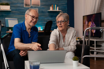 Married elder couple looking at laptop screen in living room together. Retired people using device...