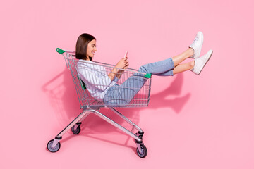 Photo of shiny cute young woman wear white sweater sitting shopping cart chatting modern gadget smiling isolated pink color background