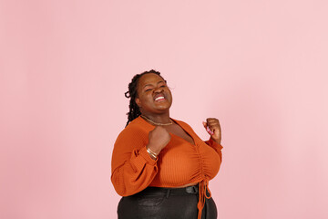 Excited young black plus size body positive woman with dreadlocks in orange top happy of big win poses for camera on pink background in studio closeup - 468122596