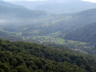 Landscape of a mountain village surrounded by green waves of beech forests under the fog hanging in the air.