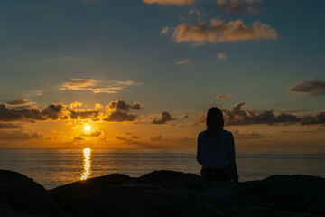 Naklejka na ściany i meble Young woman in a white shirt and jeans is sitting on huge rocks and looking at a beautiful, mesmerizing sunset over the sea, photo from the back, copy space