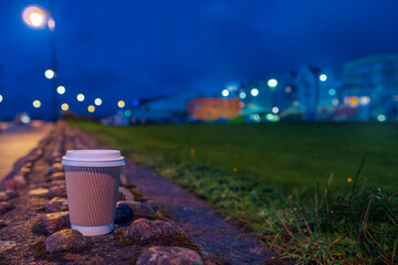 Cardboard take away cup with plastic lid and coffee or tea on a stone fence. City lights in the...