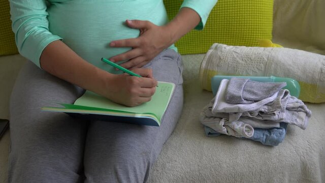 Cute Pregnant Woman writing packing list for maternity hospital With Notebook Prepares bags. Young Ledy In Pregnancy Have Fun Spend Time On Sofa At Home. Motherhood, Medicine Health and Care Concept