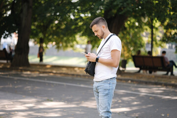 Fototapeta premium Man in white t-shirt with black shouler bag using phone outddor. Man has smart watch