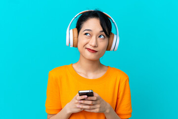 Young Vietnamese woman isolated on blue background listening music with a mobile and thinking