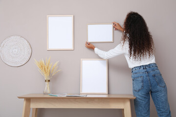 African American woman hanging empty frame on pale rose wall over table in room, back view. Mockup...