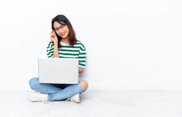 Young Vietnamese woman with a laptop sitting on the floor isolated on white wall with glasses and happy