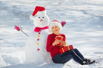 A young girl dressed in a Santa hat and a red sweater holds gifts in her hands and hugs a Snowman