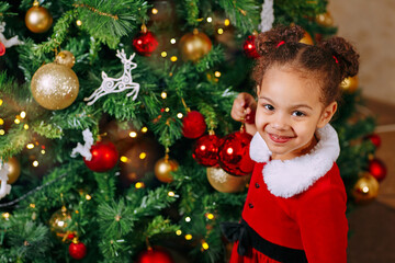 A portrait of black girl in a red dress decorates a Christmas tree with a red glass red ball at home.