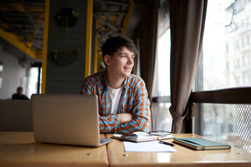 Smiling male student sitting front open laptop computer while looking away. Young man freelancer working remote  with online project on netbook