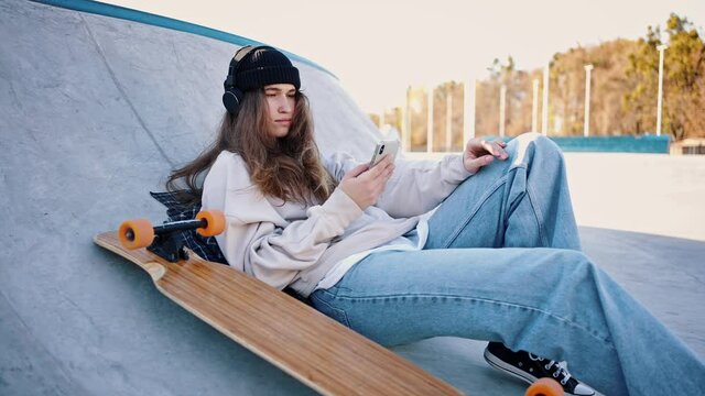 Casual dressed teenage girl lies on a springboard in a skatepark listening to music. Portrait of a girl and her longboard