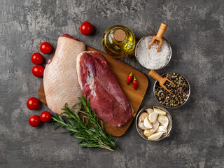 Raw duck fillet and spices for cooking Christmas and Thanksgiving festive dish. Poultry breast, cherry tomatoes, garlic, fresh rosemary, salt, pepper and olive oil over dark surface.