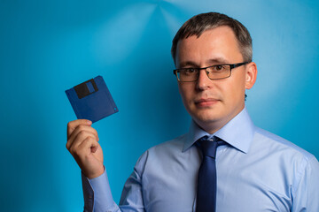A man in a blue shirt, tie and glasses showing a blue floppy disk. Retro technology concept.