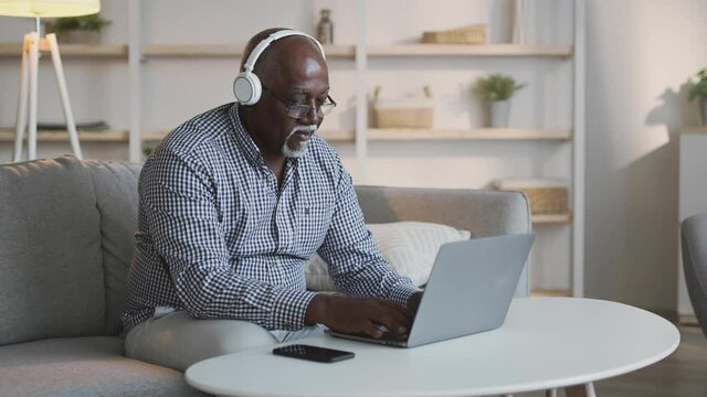Positive trendy senior african american man listening to music in wireless headphones while working on laptop at home