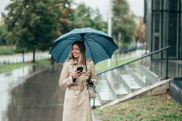 Businesswoman with umbrella using smartphone and walking down city street during rain