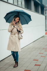 Businesswoman with umbrella using smartphone and walking down city street during rain