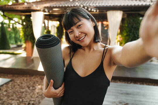Asian woman taking selfie photo while standing with yoga mat
