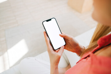 Woman holding a smartphone with a white screen mock up, resting on a sofa in living room at home.