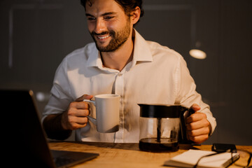 White bearded man drinking coffee while working with laptop in evening