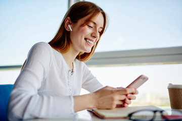 Cheerful female student in electronic earbuds enjoying mobile messaging in social networks using...