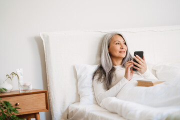 Grey asian woman using mobile phone while lying in bed after sleep