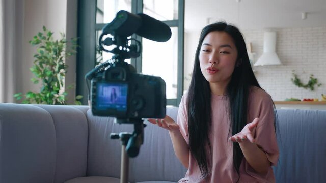 Happy young asian woman blogger filming new vlog video with professional camera mounted on tripod in kitchen at home. Blogging concept.