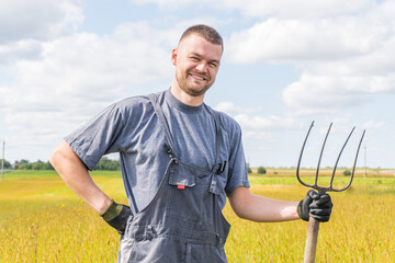 Portrait of a young smiling farmer in a field in summer. Farmer with a pitchfork, looking at...