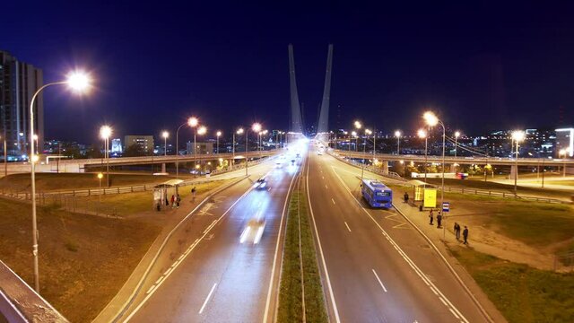 Timelapse from above of the bridge road. Illuminated Golden Bridge in Vladivostok at night, driving cars, public transport and people waiting at bus stops