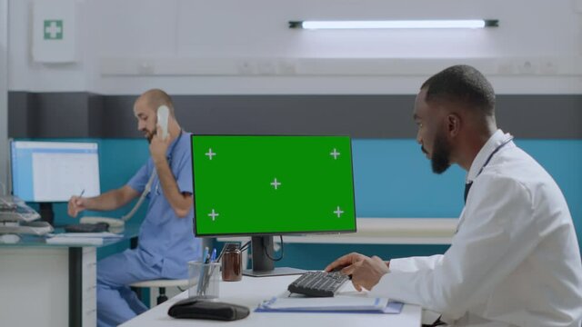 African american doctor sitting at desk in hospital office working at healthcare treatment typing medical expertise on mock up green screen chroma key computer with isolated display. Medicine concept
