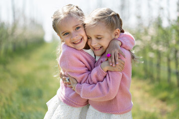 Happy two twin sisters are hugging against the background of a green blossoming apple orchard - obrazy, fototapety, plakaty
