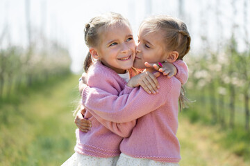 Happy twin sisters hug against the background of a green blooming Apple orchard. One sister kisses the other on the cheek
