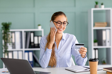 Mobile payments. Happy businesswoman holding credit card and talking on smartphone, sitting on workplace