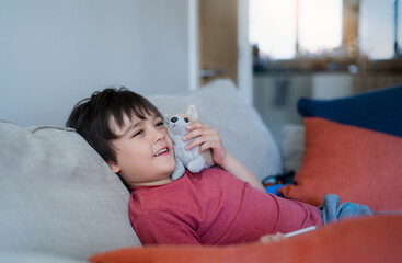 Happy young boy sitting on sofa playing with soft toy while watching cartoon on TV. Cute Child resting in living room with light shining from window on sunny day summer. Kid relaxing alone at home