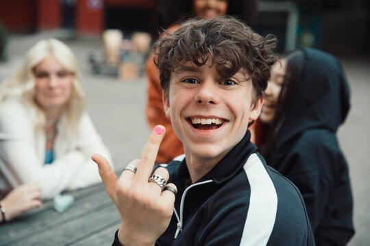 Portrait of happy teenage boy doing obscene gesture while sitting with friends