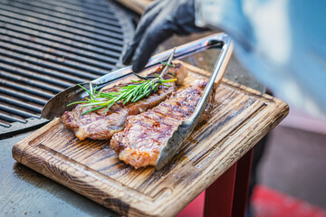 Delicious juicy Beef Steaks with rosemary and kitchen tongs on wooden cutting board, serving, cook hand