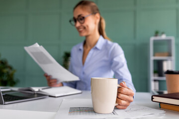 Young woman holding coffee cup and reading documents in office interior, sitting at workplace, selective focus