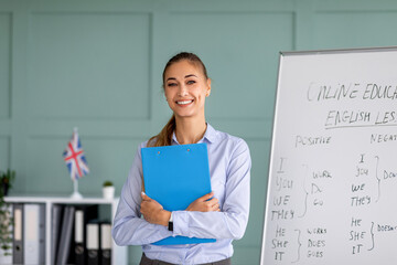 Happy teacher giving virtual class, holding clipboard and smiling at camera, standing near...