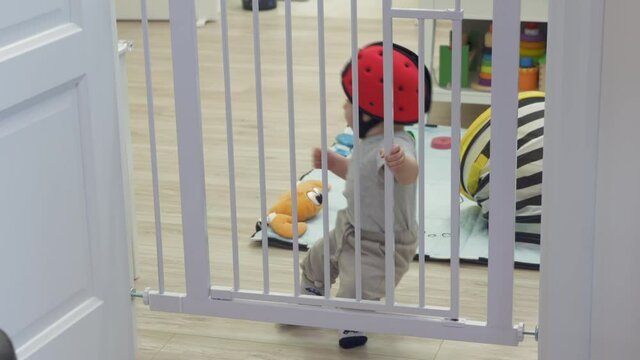 Toddler in safety helmet falling backwards and hitting his head on the floor, cute child learns to stand walk holding on to safety baby gates. Baby safety proofing at home. High quality 4k footage