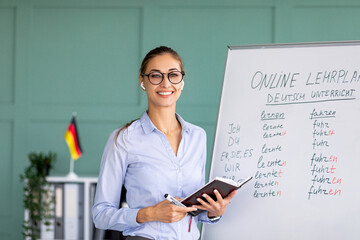 Portrait of cheerful German teacher standing near blackboard, conducting internet lesson and...