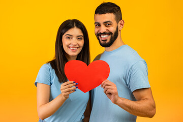 Fototapeta na wymiar Valentine's day. Loving arab spouses embracing and holding heart, smiling to camera over yellow studio background